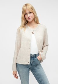 Cárdigan beige claro hecho de una tela suave, con un cuello redondo, cierre de botones y mangas largas. Llevado sobre una blusa blanca y jeans azules.