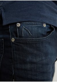 Tmavě modré džíny z denimu s přední kapsou, na které je malý prošitý detail. Je viditelná hladká textura a stříbrné nýty.