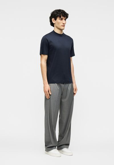 Νέος άνδρας στέκεται φορώντας μπλε ναυτικό κοντομάνικο πουκάμισο, φαρδιά γκρι παντελόνια και λευκά αθλητικά παπούτσια μπροστά σε απλό φωτεινό φόντο.