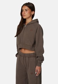 Sweat à capuche court en tissu doux avec fermeture éclair, poignets élastiques et logo rond sur la poitrine, associé à un pantalon de jogging assorti.