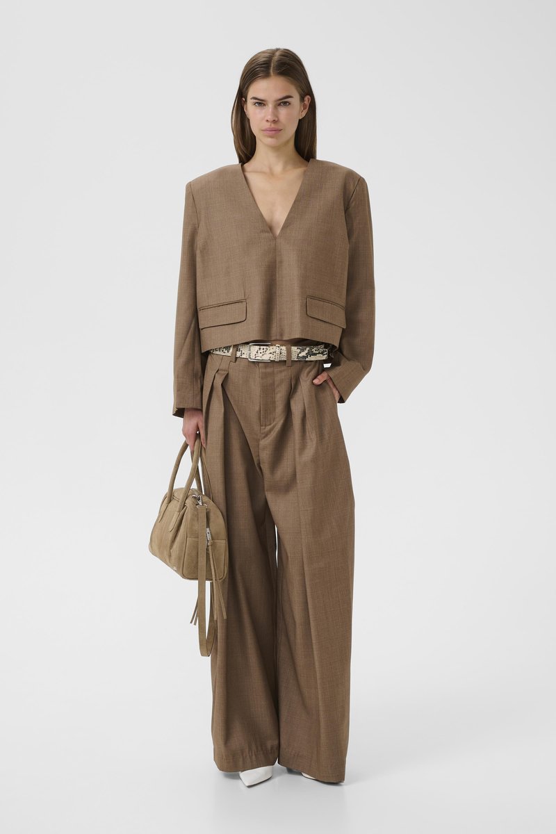 Femme portant un pantalon large marron et un blazer court à col en V, tenant un sac à main beige, avec une ceinture à motifs et des chaussures blanches.