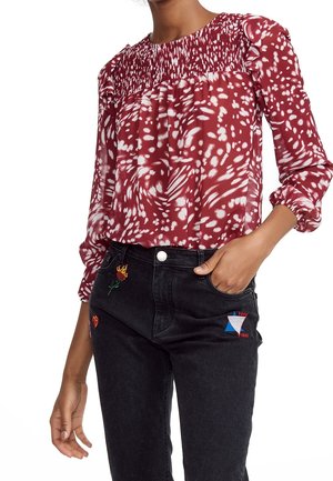 Femme portant un chemisier à manches longues rouge et blanc à motifs avec un plissé sur la poitrine, associé à un jean noir avec des patches brodés colorés.