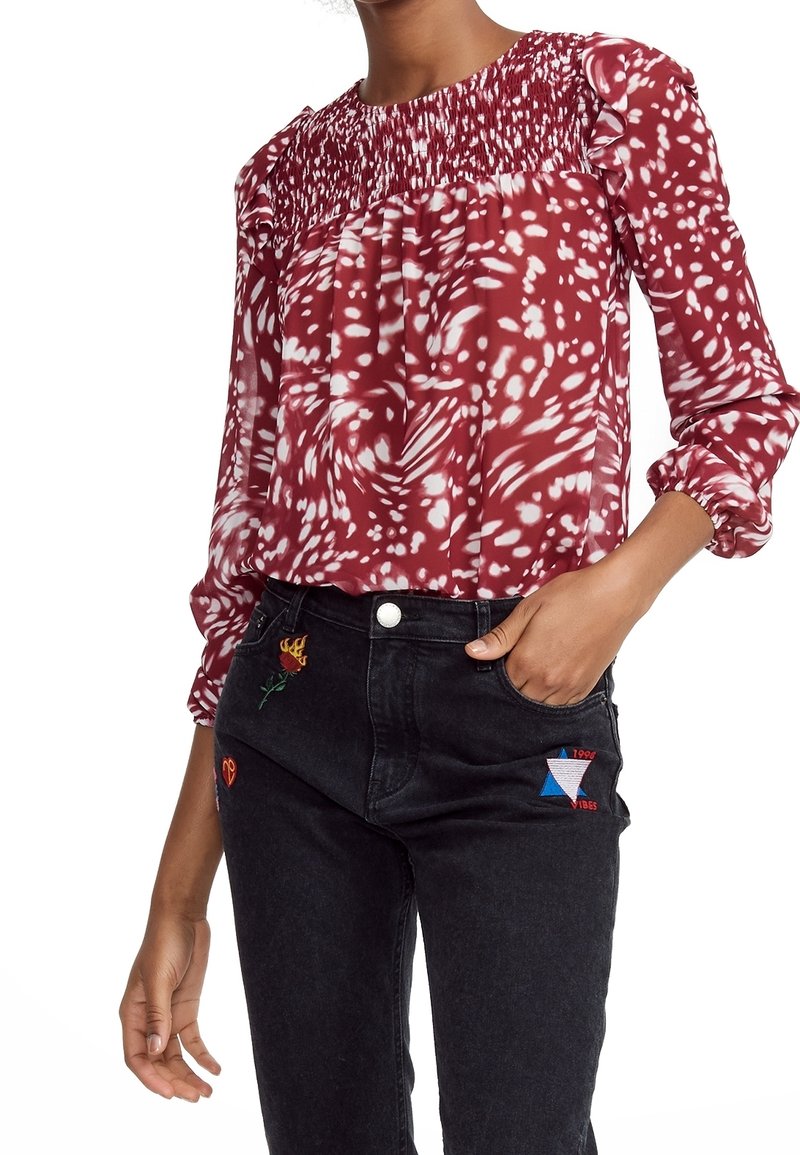 Femme portant un chemisier à manches longues rouge et blanc à motifs avec un plissé sur la poitrine, associé à un jean noir avec des patches brodés colorés.