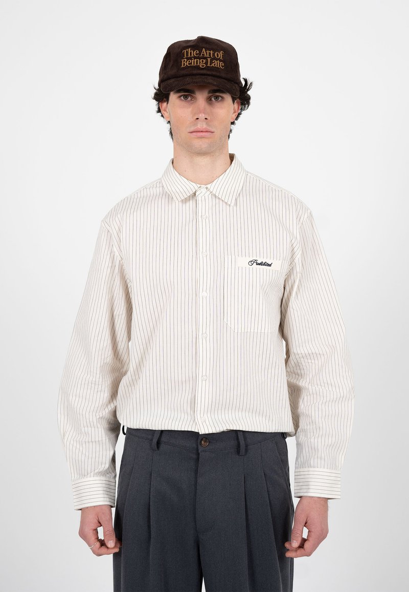 Homme portant une casquette marron avec le texte « The Art of Being Late », une chemise à rayures blanches boutonnée et un pantalon plissé foncé sur un fond blanc.