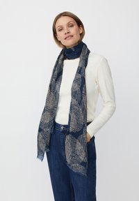 Marineblå tørklæde med et geometrisk mønster i beige, frynsede kanter, lavet af et blødt, struktureret stof, båret over en lysefarvet top.
