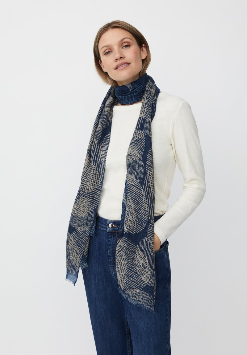 Marineblå tørklæde med et geometrisk mønster i beige, frynsede kanter, lavet af et blødt, struktureret stof, båret over en lysefarvet top.