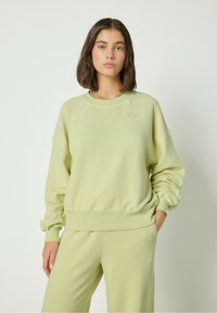 Gaiši zaļa džemperis, kas izgatavots no mīkstas materiāla, ar ribotām piedurknēm, apaļu dekoltē un brīvu piegriezumu. Saskaņots ar atbilstošajām biksēm.