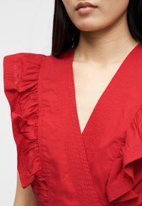 Γυναίκα που φοράει κόκκινο φόρεμα με βαθύ V λαιμόκοψη και βολάν σε κοντά μανίκια, εμφανιζόμενη από τους ώμους έως το άνω μέρος του στήθους.