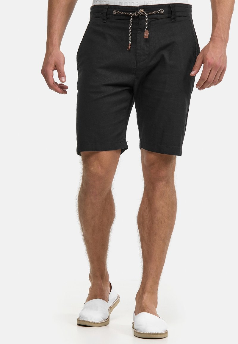 Homme portant un short noir jusqu'aux genoux avec une taille à cordon et des chaussures blanches sans lacets, debout devant un fond uni.