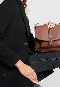 Henkilö, jolla on päällään musta takki ja tummat farkut, pitää ruskeaa nahkabagia vaaleata taustaa vasten.