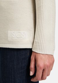 Beige Rippstrickpullover mit strukturierter Oberfläche und einem Etikett mit der Aufschrift "SCSY" in einer kontrastierenden hellen Farbe. Getragen über dunklen Jeans.