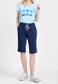 Frau trägt ein hellblaues GAP T-Shirt, marineblaue knielange Shorts mit Kordelzug und weiße Turnschuhe, steht mit den Händen in den Taschen.