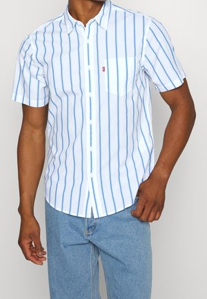 Hombre que lleva una camisa blanca de manga corta con botón, con rayas verticales azules y bolsillo en el pecho, combinada con jeans azul claro.