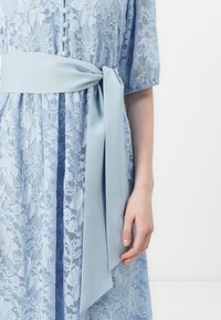 Lyseblå blondekjole med blomstermønster, korte ermer og et matchende satengbelte knyttet i livet. Knapper pryder fronten.