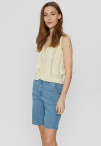 Brede gebreide cardigan in lichtbeige met verticale strepen, voorzien van een V-hals en knopen aan de voorkant. Gecombineerd met blauwe denim shorts.