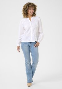 Camicia bianca a bottoni con dettagli in pizzo, maniche lunghe e colletto, abbinata a jeans a zampa d'elefante blu chiaro e calzature beige.