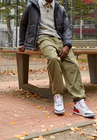 Vadderad svart jacka, grå polo skjorta och löst sittande olivgröna byxor. Vita och röda sneakers med grå accenter. Sitter på en bänk bland löv.