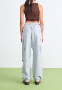 Person with braided hair wears brown sleeveless top, loose light gray cargo pants with back and side pockets, and black platform shoes on green floor.