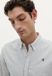 Camicia a righe in tessuto leggero con righe verticali blu navy e bianche. Presenta un colletto e un'emblema di teschio sul lato sinistro del petto.