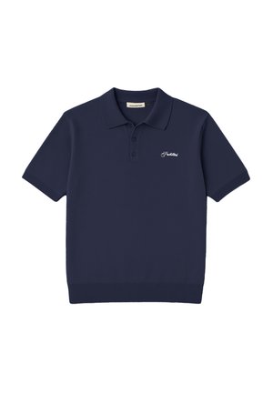 Ung mand iført en navy poloshirt, grå fløjlsbukser med fine striber, mørkegrøn kasket med orange logo, holder brune handsker i venstre hånd mod hvid baggrund.