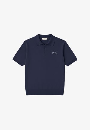 Jeune homme portant un polo bleu marine, un pantalon gris à fines rayures, une casquette vert foncé avec un logo orange, tenant des gants marron dans la main gauche sur fond blanc.