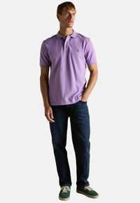 Violeta polo krekls no kokvilnas ar smalku tekstūru, ar apkakli un mazu zaļu logotipu. Kombinēts ar tumšiem džinsiem un zaļiem sporta apaviem.