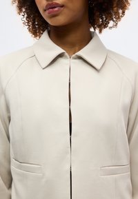 Γυναίκα με σγουρά μαλλιά φοράει μπεζ μπουφάν με μυτερό γιακά και κούμπωμα με άγκιστρο και ματιές, μερικώς κουμπωμένο πάνω από μαύρο τοπ.