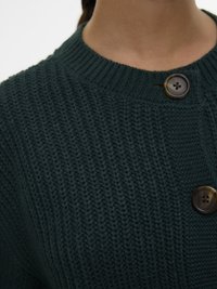Close-up de camisola de malha verde escura com gola canelada e dois grandes botões castanhos perto do decote numa pessoa.