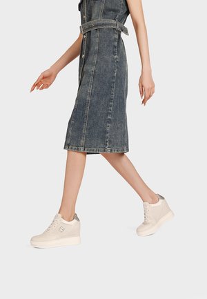 Donna che cammina indossando un vestito di denim sbiadito di lunghezza media con bottoni e scarpe da ginnastica bianche con zeppa e dettagli argentati sul tacco.