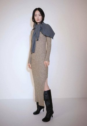 Robe en tricot beige avec une fente sur le côté, accompagnée d'une écharpe gris foncé drapée sur les épaules, et de bottes à talons noires. Fond clair et uni.