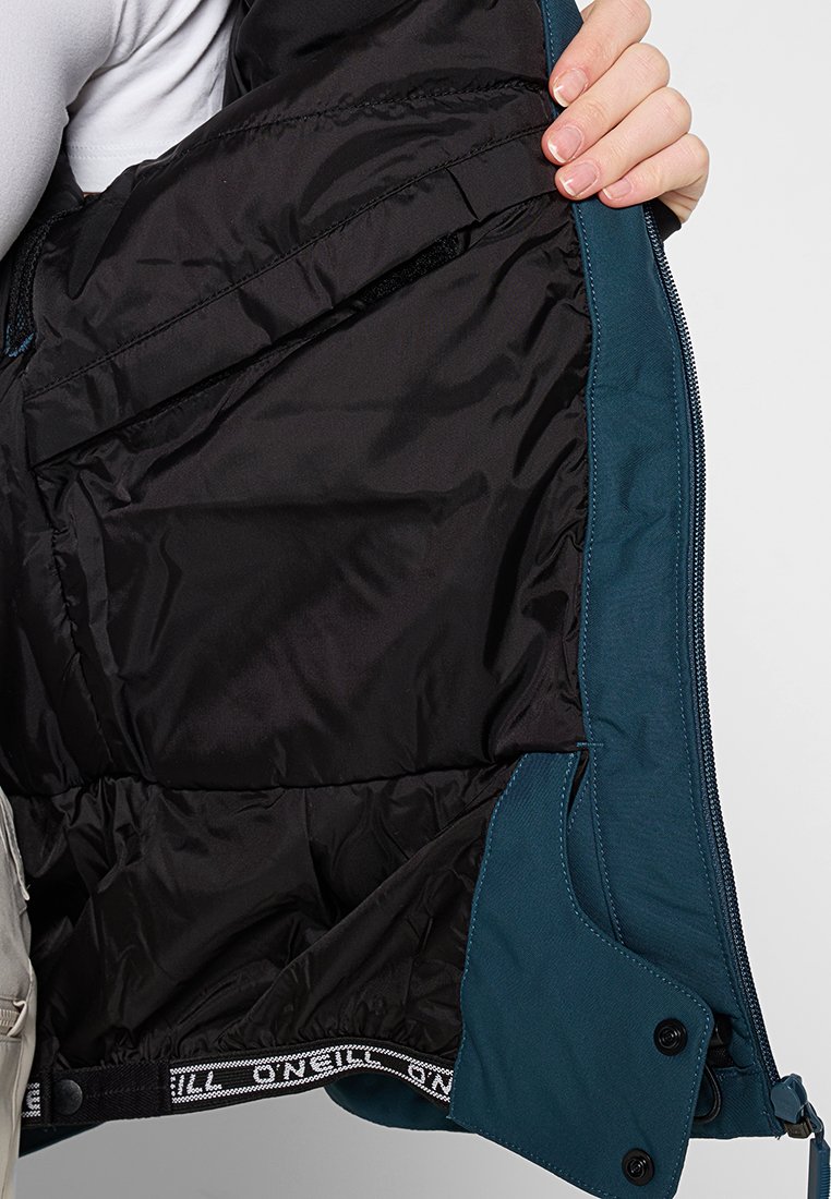 Eine Hand hält eine offene türkisfarbene Jacke, die das schwarze gepolsterte Innenfutter und einen verstellbaren Riemen mit dem Text "O'Neill" zeigt.