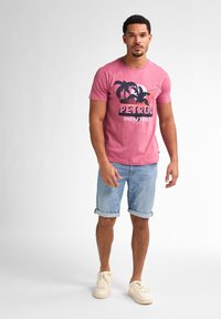 T-shirt rosa in cotone con grafica di una palma nera, abbinata a pantaloni corti in denim azzurro arrotolati e sneakers color crema.