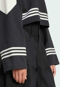 Černá zkrácená mikina s bílými pruhy na rukávech. Ruka modelky spočívá na tmavých kalhotách s texturovaným povrchem.