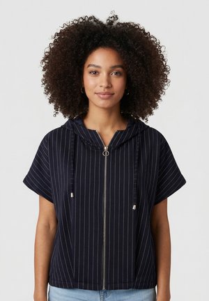 Mujer con cabello rizado que lleva una sudadera con capucha de manga corta azul marino con rayas blancas y jeans azul claro contra un fondo liso.