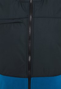 Chaqueta acolchada con una cremallera negra vertical, sección superior negra y sección inferior de forro polar azul. Cuenta con un bolsillo en el pecho y una textura suave.