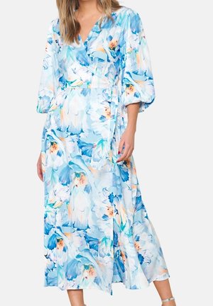 Mujer con un vestido largo, fluido, azul pastel con estampado floral, manga abullonada tres cuartos y cintura anudada.