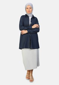 Denim button-up shirt in donkerblauw, gedragen over een lange lichtgrijze rok. Het model draagt een lichtgrijze hijab en beige sandaaltjes met bandjes.