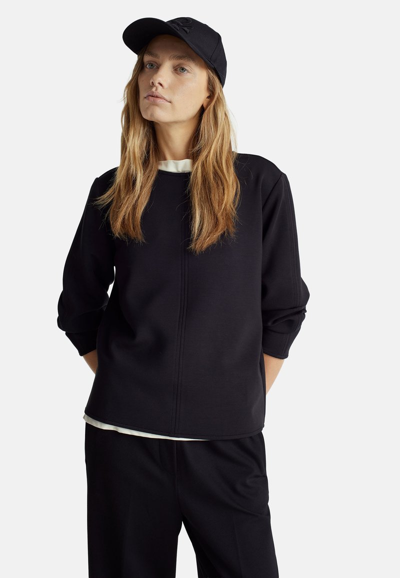 Schwarzer Sweatshirt mit langen Ärmeln, Rundhalsausschnitt und subtilen Nahtdetails, kombiniert mit hochtaillierten dunklen Hosen und einer schwarzen Kappe.