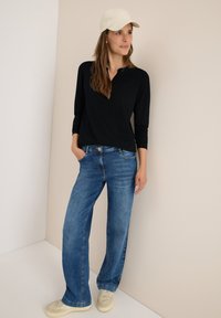 Frau in einem schwarzen Langarmshirt, blauen Jeans, beigem Hut und cremfarbenen Turnschuhen, die an einer Ecke wand steht.