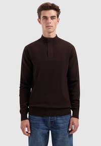 Junger Mann in einem dunkelbraunen Langarmpullover mit hohem Kragen und blauen Jeans, der vor einem einfarbigen hellen Hintergrund steht.