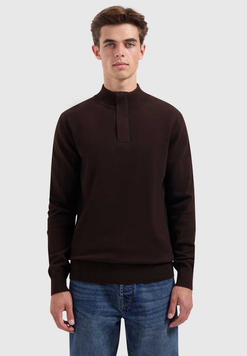 Junger Mann in einem dunkelbraunen Langarmpullover mit hohem Kragen und blauen Jeans, der vor einem einfarbigen hellen Hintergrund steht.