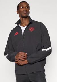 Μαύρο αδιάβροχο μπουφάν με κόκκινο λογότυπο της Manchester United, ψηλό γιακά, φερμουάρ κλεισίματος, ελαστικές μανσέτες και λευκές ρίγες στα μανίκια.