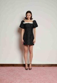 Černé krátké šaty s nafouklými rukávy a bílým detailem u výstřihu, s přiléhajícím střihem a hladkou texturou. Nošeny s leopardím vzorem na podpatcích.