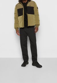 Man som bär en olivgrön och svart pufferjacka, svart skjorta, mörkgrå byxor och svarta sneakers som står mot en vit bakgrund.