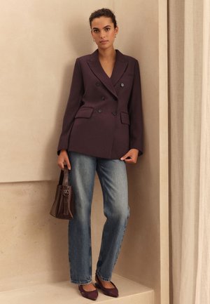 Femme portant un blazer croisé violet foncé, un jean bleu, des ballerines pointues assorties, tenant un sac à main marron foncé, debout contre un mur beige.