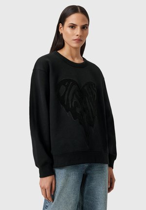 Felpa nera con un design a cuore testurizzato. Presenta un colletto rotondo e maniche lunghe, abbinata a jeans in denim blu chiaro. Tessuto morbido al tatto.