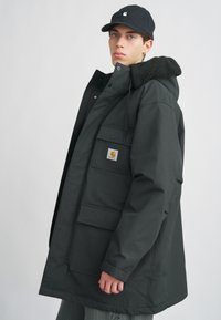 Must mustaga varrukat, millel on karvane kaelus, mitmed ees olevad taskud ja logo silt. Sile tekstuur, põlvepikkune disain ja vabalt langev lõige.