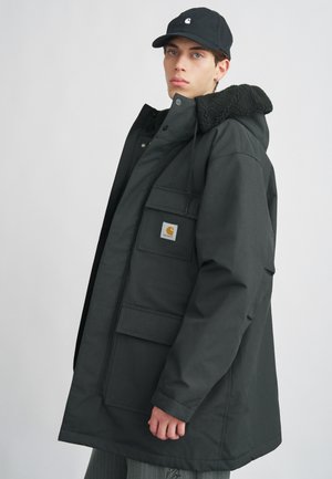 Schwarze isolierte Jacke mit fellgefütterter Kapuze, mehreren Fronttaschen und einem Logo-Patch. Glatte Textur, knielanges Design und lässige Passform.