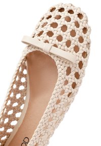 Bailarina beige tejida con un diseño abierto, que presenta un pequeño lazo en la puntera y una superficie texturizada. Forro interior suave visible.