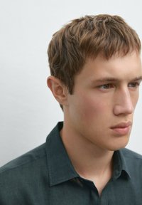 Dark green collared shirt with a textured fabric, worn by a male model with short, light brown hair, facing slightly to the left.